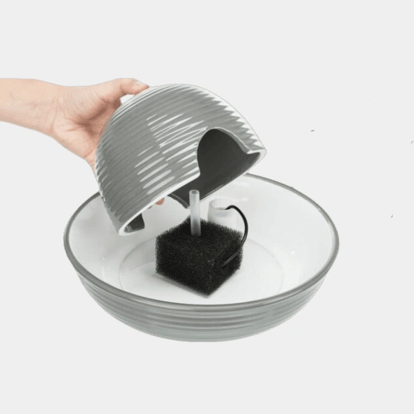 Mano sosteniendo la tapa de un bebedero automático para perros, mostrando el filtro y la bomba internos, destacando la facilidad de limpieza y mantenimiento