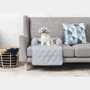 Perro blanco descansando sobre un protector de sofá para perros acolchado en un sofá gris, con cojines decorativos y una cesta de ropa al lado