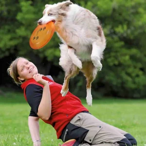Perro saltando y atrapando un frisbee canino naranja en el parque, mientras su dueña lo anima