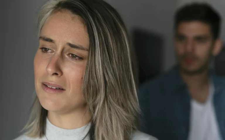 Mujer con expresión de tristeza mientras su pareja se encuentra desenfocada en el fondo, representando el impacto emocional de una separación o divorcio